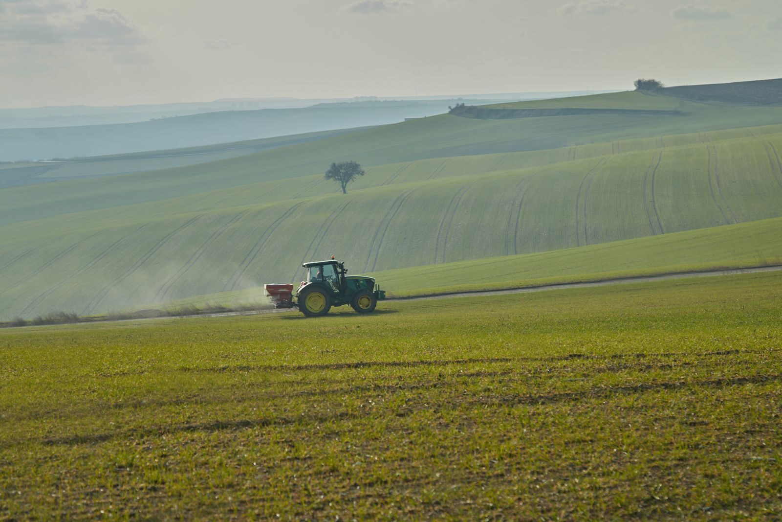 A tractor pulling a spreader across a field, with dust rising behind it