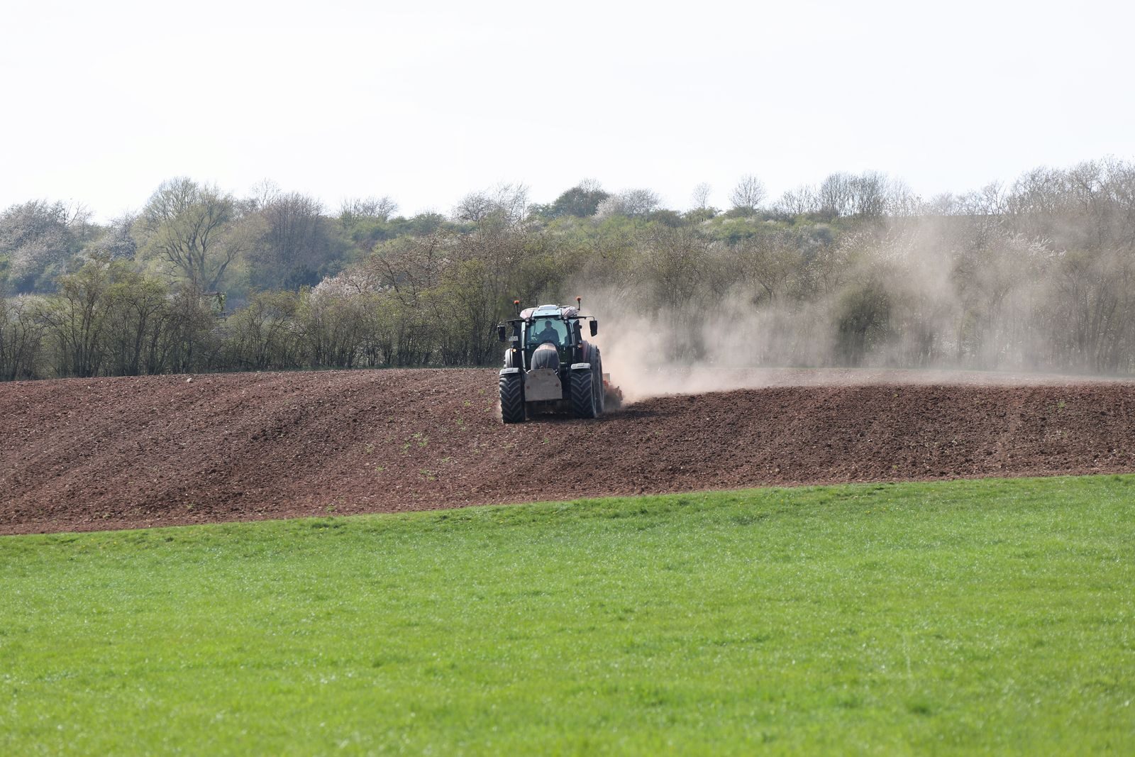 A tractor spreading fine material across a bare field, with dust rising behind it